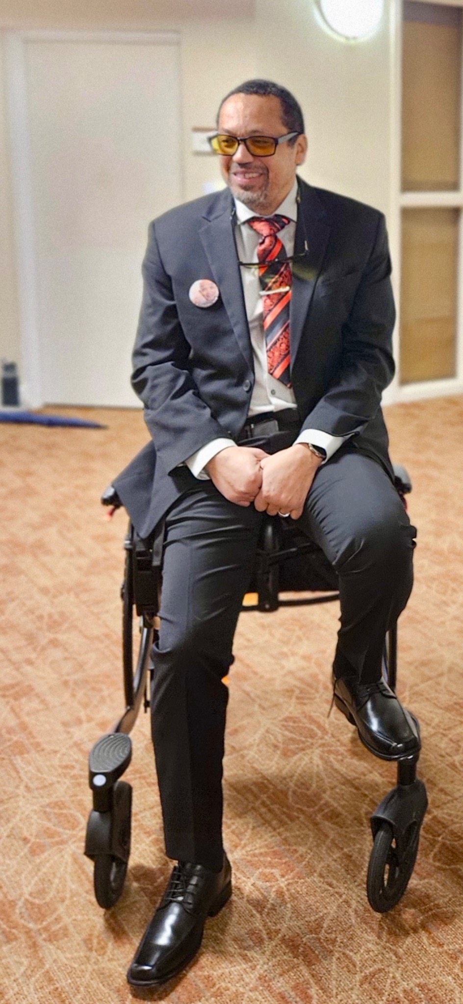 Mike smiling in a dark suit and red tie while seated in his wheelchair